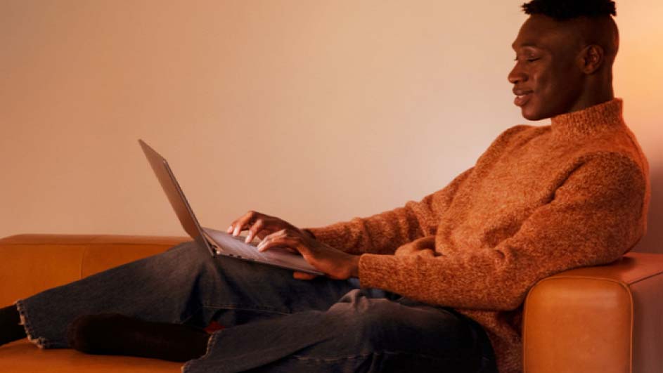 Un homme souriant assis confortablement dans un canapé orange, utilise un ordinateur portable.