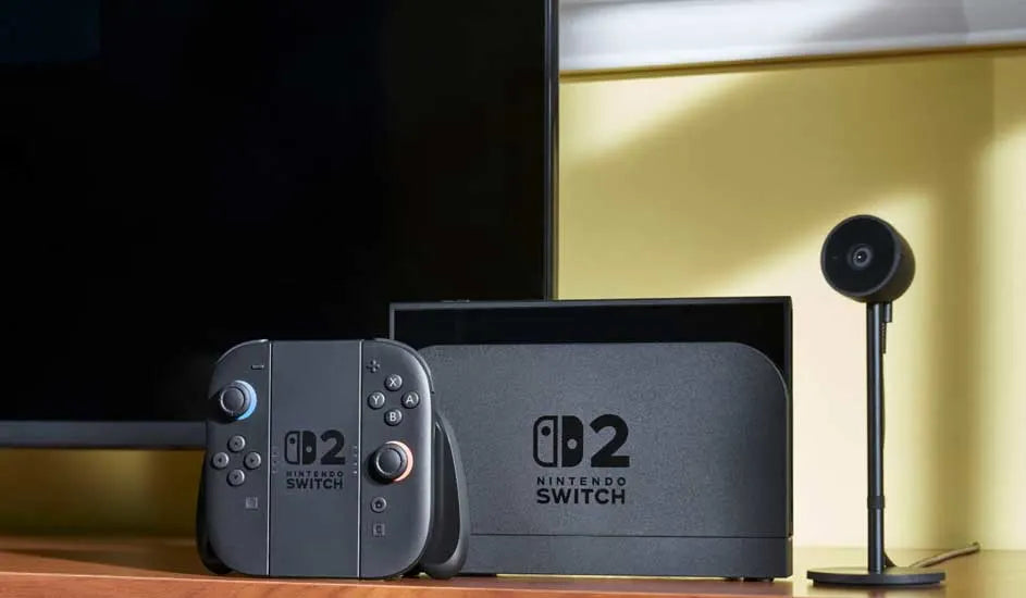 Une Nintendo Switch 2 dans sa station de charge avec un Joy-Con posé sur un meuble à côté d'une TV et d'une caméra.