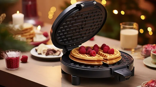 Un gaufrier ouvert avec gaufres garnies de framboise, sur une table décorée de bougies à l'ambiance chaleureuse.