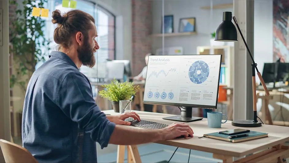 Un homme barbu travaille sur son ordinateur dans un bureau, des graphiques d'analyse sont affichés sur l'écran.
