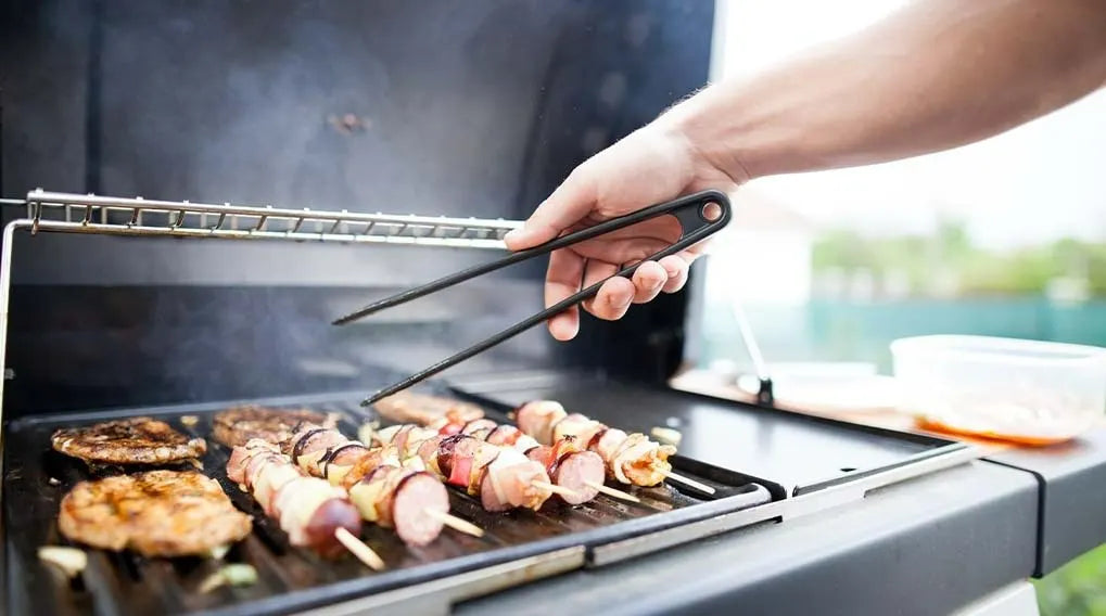 Une main tient une pince au dessus d'un barbecue sur lequel cuisent de la viande et des brochettes.