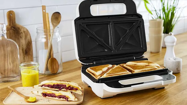Un appareil à croque-monsieur ouvert avec sandwichs grillés, près d'un croque-monsieur et d'un pot de sauce sur une table en bois.
