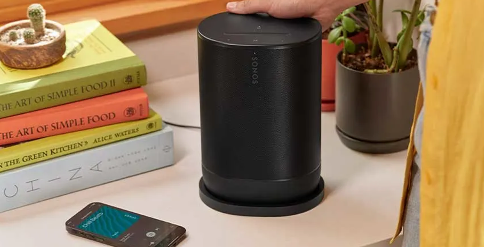 Une enceinte Sonos noire sur la base de charge à côté de livres, d'une plante et d'un smartphone.