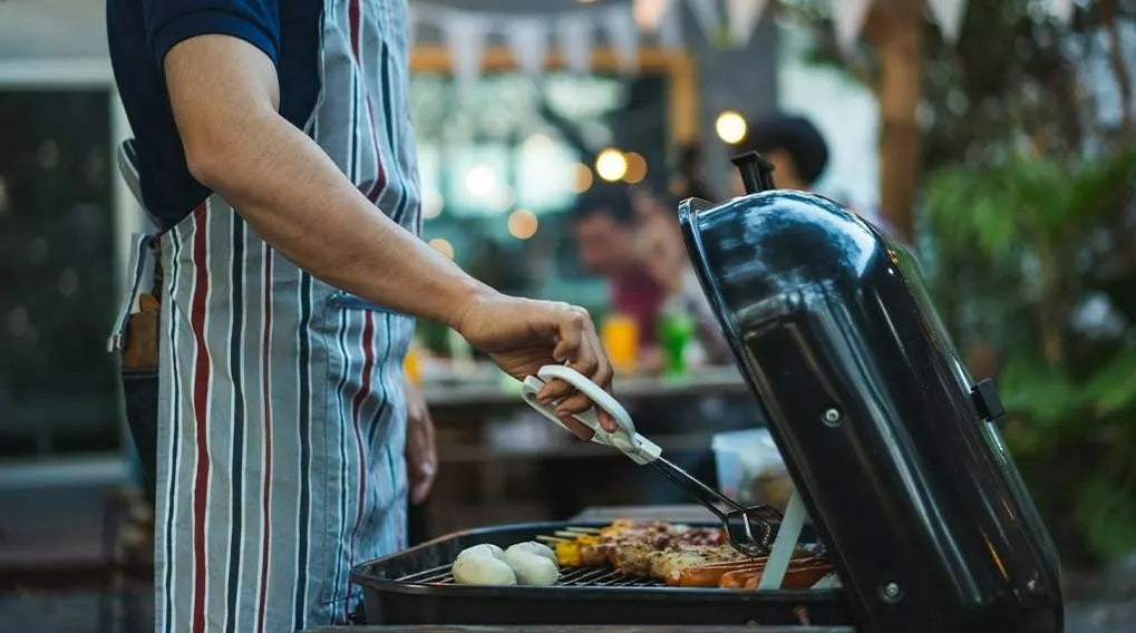 Une personne utilise une pice pour cuire des brochettes et des saucisses sur un barbecue dans un jardin.