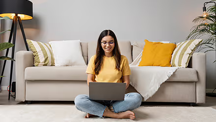 Une femme souriante, assise en tailleur devant un canapé, utilise un ordinateur portable. 