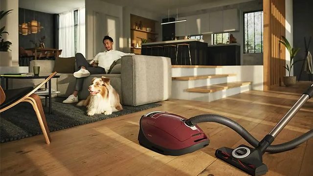 Un aspirateur de couleur bordeaux au premier plan pendant qu'un jeune homme et son chien se relaxe sur un canapé au second plan.