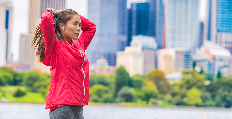 Une femme en tenue de sport rouges avec des écouteurs aux oreilles, se prépare à courir devant la ville.