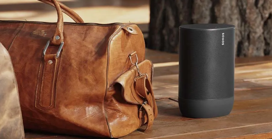 Une enceinte Sonos noire posé sur du parquet , près d'un petit sac de voyage en cuir.