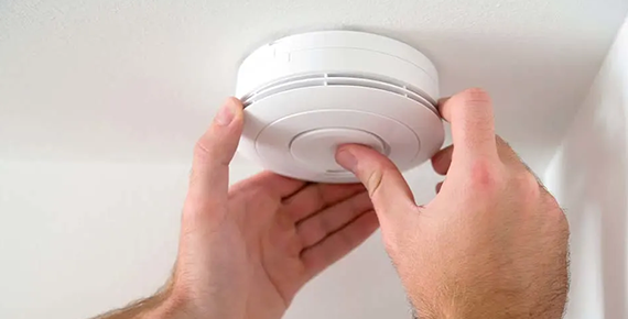 Deux mains achève de fixer un détecteur de fumée blanc au plafond.