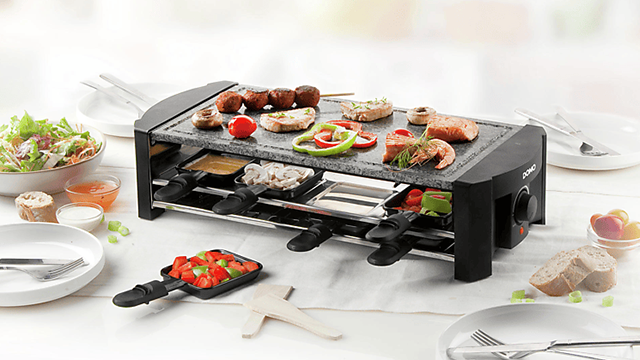 Un appareil a pierrade-raclette noir Domo avec crevettes, brochettes, légumes, et poêlons sur une table dressée.