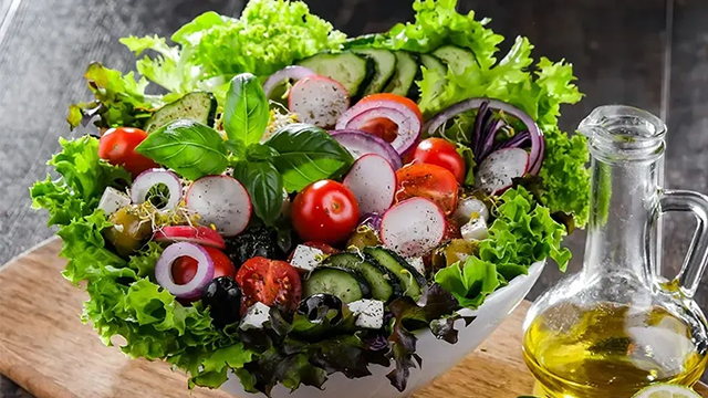 Une salade composée de laitue, tomates, concombre, tomates et oignon dans un saladie à côté d'une bouteille d'huile d'olives.