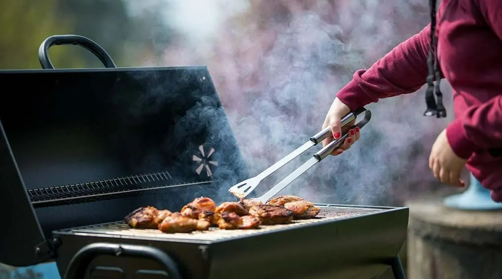 Une personne retourne de la viande avec une pince sur barbecue fumant en extérieur.