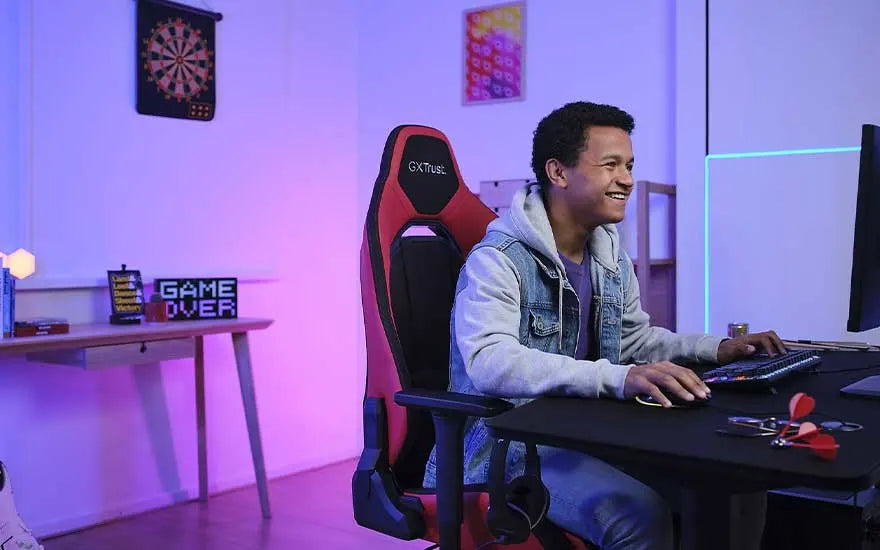 Un joueur souriant installé dans une chaise gaming, devant une installation comprenant, un clavier, une souris, un moniteur et un bureau dans une pièce baignée d'une lumière violette.