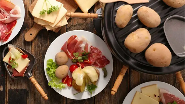 Une table vue du dessus avec un appareil à raclette des assiettes contenant des pommes de terre, des légumes et de la charcuterie.