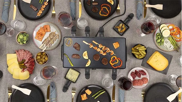 Une table conviviale vue de dessus avec raclette-grill, brochettes, fromages, charcuterie, légumes et assiettes noires.