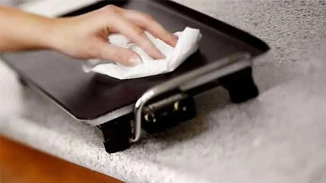Une main essuie la plaque d'un grill avec un chiffon blanc sur un plan de travail.