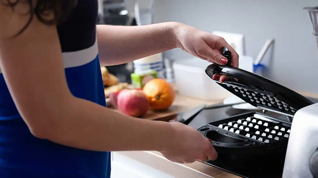 Une personne ouvre un gaufrier sur un plan de travail avec des fruits en arrière plan.