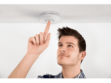 Charger l&#39;image dans la galerie, Un homme appuie sur un bouton d&#39;un détecteur de fumée blanc monté sur un plafond blanc.
