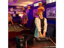 Charger l&#39;image dans la galerie, Femme DJ avec un casque et une table de mixage, avec un ordinateur portable et une enceinte, dans un club.
