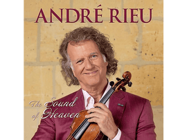 André Rieu tient un violon, souriant. Il porte une chemise rose et une veste rouge. Derrière lui, un mur de pierre.