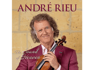 André Rieu tient un violon, souriant. Il porte une chemise rose et une veste rouge. Derrière lui, un mur de pierre.