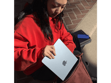 Charger l&#39;image dans la galerie, Une femme en sweat-shirt rouge tient un ordinateur portable bleu clair avec un logo Apple.
