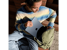 Charger l&#39;image dans la galerie, Une personne assise, ouvrant un sac à dos vert, avec un ordinateur portable bleu à la main, assise sur des briques.
