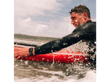 Charger l&#39;image dans la galerie, Homme sur une planche de surf rouge dans l&#39;océan. Il porte une combinaison et des écouteurs.
