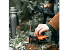Charger l&#39;image dans la galerie, Mains d&#39;une personne utilisant un drone avec une manette et un téléphone, sur une bûche, en pleine nature.
