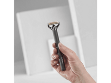 Charger l&#39;image dans la galerie, Une main tient un rasoir BaByliss noir et doré. Le fond présente des formes géométriques blanches.
