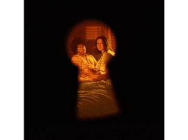 Une vue à travers un trou de serrure montre deux personnes dans une pièce. La scène est éclairée en orange. Ils sont assis sur un lit.