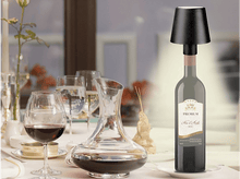 Charger l&#39;image dans la galerie, Bouteille de vin avec lampe sur une table. Verres et carafe présents. Cadre de restaurant.
