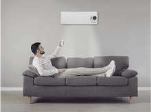 Charger l&#39;image dans la galerie, Homme sur un canapé gris utilisant une télécommande pour contrôler un climatiseur. Climatiseur sur le mur gris. Pièce blanche.
