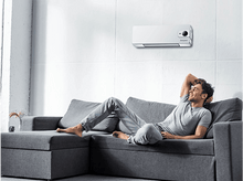 Charger l&#39;image dans la galerie, Homme sur un canapé gris, se relaxant et souriant, avec un climatiseur blanc sur le mur derrière lui.
