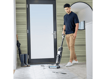 Charger l&#39;image dans la galerie, Un homme passe l&#39;aspirateur. Porte, miroir, bottes en arrière-plan. Mur en bois clair.
