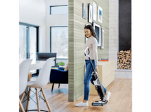 Charger l&#39;image dans la galerie, Femme passant l&#39;aspirateur sur un sol en bois dans un salon moderne avec des meubles blancs.
