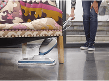Charger l&#39;image dans la galerie, Un balai vapeur nettoie le sol à côté d&#39;un canapé à motifs. Une personne se tient à proximité.

