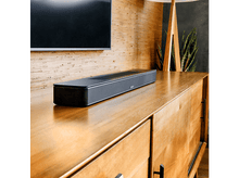 Charger l&#39;image dans la galerie, Une barre de son noire sur un meuble en bois dans une pièce avec une TV et une plante.
