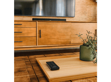 Charger l&#39;image dans la galerie, Un meuble en bois avec une TV, une barre de son noire, un plateau avec une télécommande et un vase vert avec des plantes.
