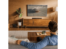 Charger l&#39;image dans la galerie, Scène de salon. Une personne sur un canapé regarde la télévision au-dessus d&#39;un meuble en bois. Barre de son visible.
