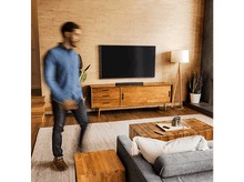 Charger l&#39;image dans la galerie, Scène de salon : TV sur meuble en bois, lampe, canapé, et personne marchant au premier plan.
