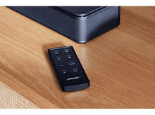 Charger l&#39;image dans la galerie, Télécommande noire Bose sur une table en bois, avec une barre de son noire en arrière-plan.
