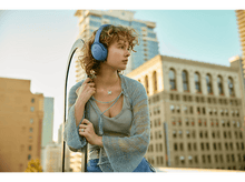 Charger l&#39;image dans la galerie, Femme avec casque bleu regarde ailleurs, fond urbain. Bâtiments visibles. Journée ensoleillée.
