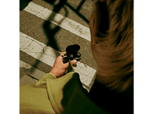 Charger l&#39;image dans la galerie, Personne tenant un étui d&#39;écouteurs noirs, avec des écouteurs visibles. Ils sont dans un lieu public.
