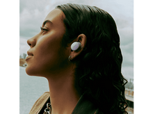 Charger l&#39;image dans la galerie, Une femme portant un écouteur lavande, souriante. Elle a des cheveux bouclés foncés et se trouve devant une vue floue de la ville.
