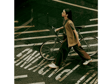 Charger l&#39;image dans la galerie, Femme avec un casque marchant à vélo. La scène est une rue avec un pavé mouillé.
