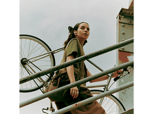 Charger l&#39;image dans la galerie, Une femme avec des écouteurs montant des escaliers avec un vélo sur l&#39;épaule, dans un cadre urbain.
