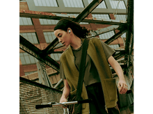 Charger l&#39;image dans la galerie, Femme portant des écouteurs et faisant du vélo sur un pont, avec un gilet et une veste verts.
