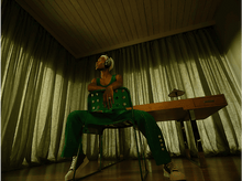 Charger l&#39;image dans la galerie, Personne assise sur une chaise, portant un casque, dans une pièce avec des rideaux verts et un bureau en bois.
