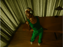 Charger l&#39;image dans la galerie, Personne portant un casque, vêtue de vert, posée sur un bureau en bois, près de rideaux verts.
